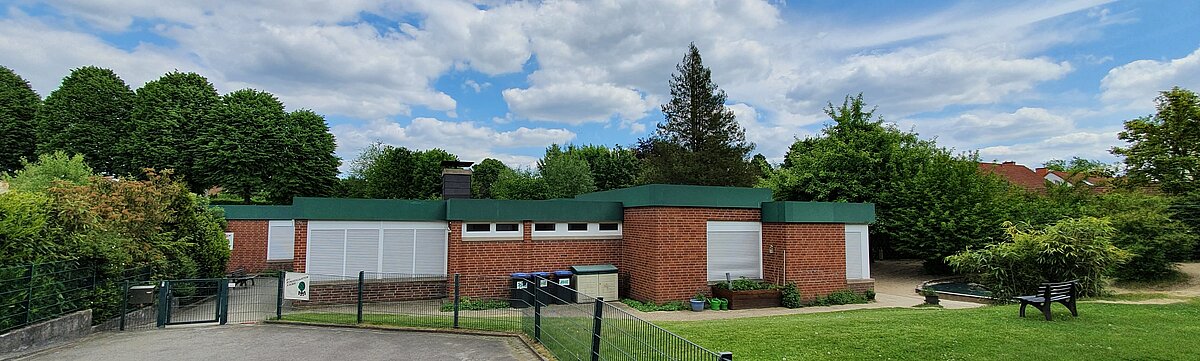 Foto: Gebäude des Kindergartens, ein eingeschossiges Flachdach-Gebäude umrahmt von einem eingezäunten Garten mit Wiesen, Bänken und Sträuchern