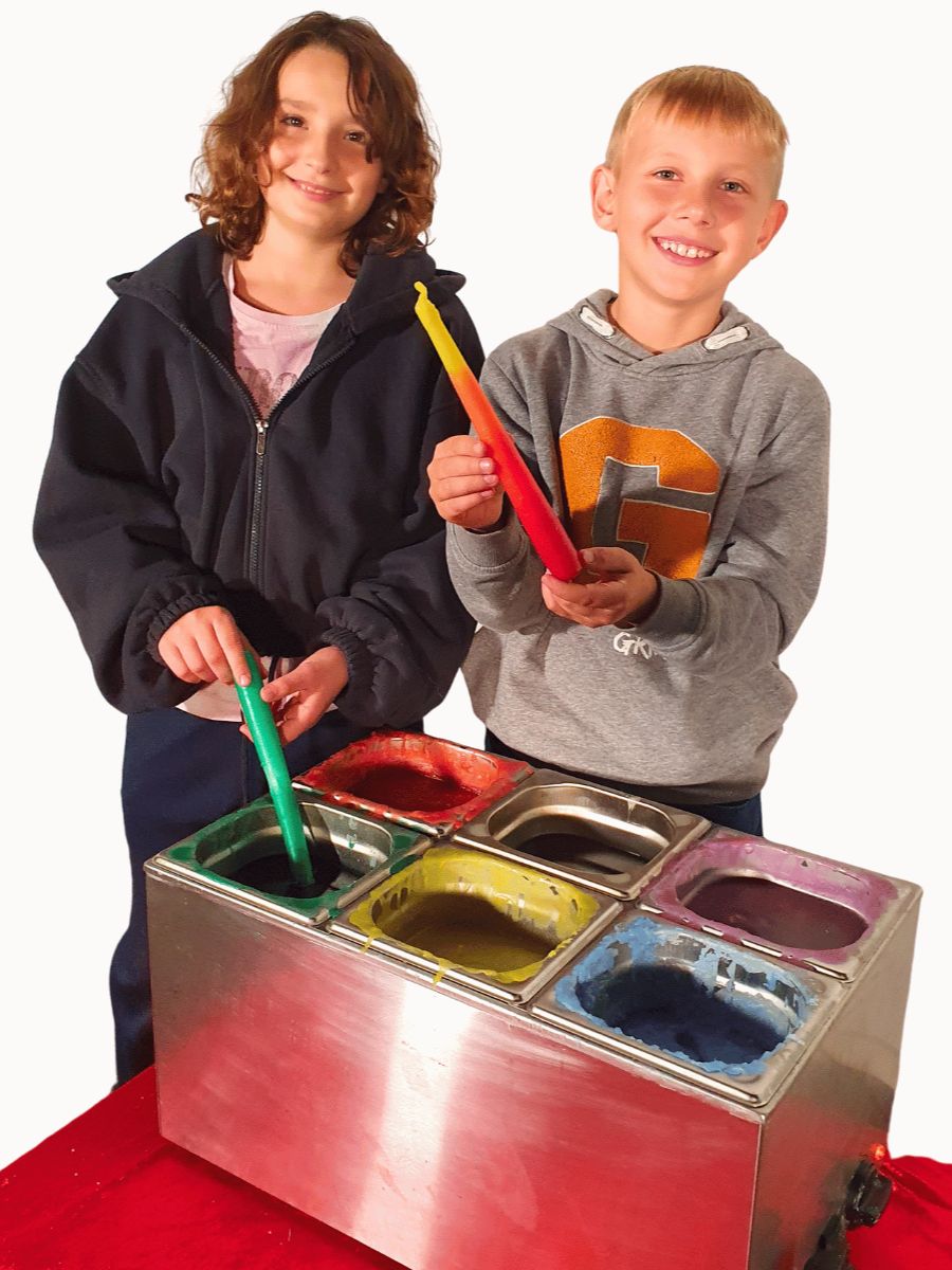 Auf dem Bild zu sehen sind zwei Kinder. Sie haben gerade eine Kerze in das Wachs getaucht. Sie zeigen ihre Kerzen in die Kamera. Vor ihnen steht das Kerzenziehgerät. Hier können die Kinder zwischen sechs verschiedenen Farben wählen: Rot, Gelb, Grün, Blau, Lila und Weiß.