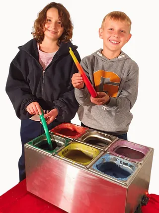 Auf dem Bild zu sehen sind zwei Kinder. Sie haben gerade eine Kerze in das Wachs getaucht. Sie zeigen ihre Kerzen in die Kamera. Vor ihnen steht das Kerzenziehgerät. Hier können die Kinder zwischen sechs verschiedenen Farben wählen: Rot, Gelb, Grün, Blau, Lila und Weiß.
