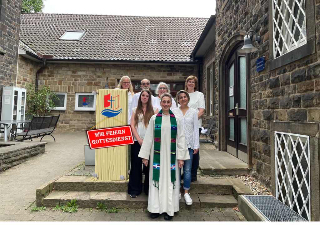 Foto vor dem Eingang zum Gemeindehaus in Heven. Das Presbyterium der Ev.Trinitatis-Kirchengemeinde Witten. Vorne in weißem Talar: Pfarrerin Heike Bundt. Dahinter Karolina Walz und Marzieh Suleighmanichli. In der dritten Reihe stehen Maike Hainsen, Udo Barthold, Brigitte Steinhoff und Ruth Tennié. Alle Presbyter*innen tragen weiße Hemden, Blusen oder T-Shirts.