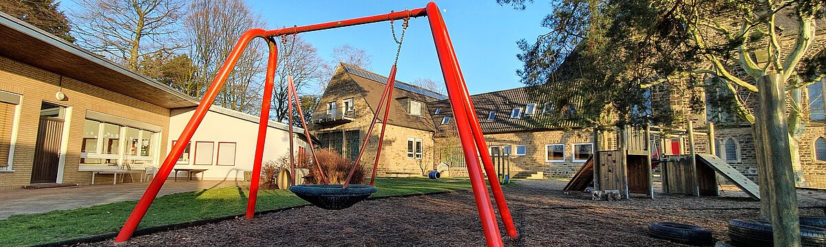Foto: Spielplatz des Kindergartens Vogelnest hinter der Kirche am Steinhügel. Ein rotes Schaukelgerüst, Sandkasten, Klettergeräte, im Hintergrund das Gemeindehaus und die Kindergartengebäude
