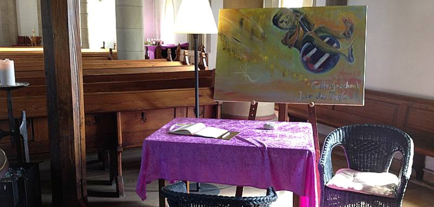 Foto: Eingangsbereich in der Kirche, hinter den letzten Sitzreihen: Ein kleiner Tisch mit lila Tischtuch, daneben ein Rattanstuhl. Im Hintergrund eine STehlampe und ein gemaltes Bild einer schaukelnden Figur, aufgestellt auf einer Staffelei