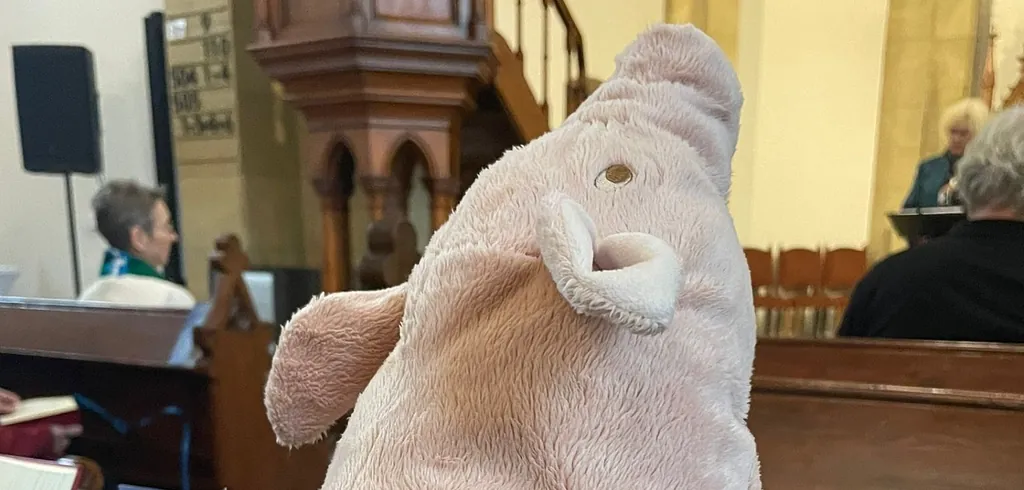 Foto: Ein rosa Plüsch-Schwein sitzt in der Kirchenbank und schaut Richtung Altar. Die Pfarrerin sitzt in der ersten Reihe. Jemand steht vorn und spricht.
