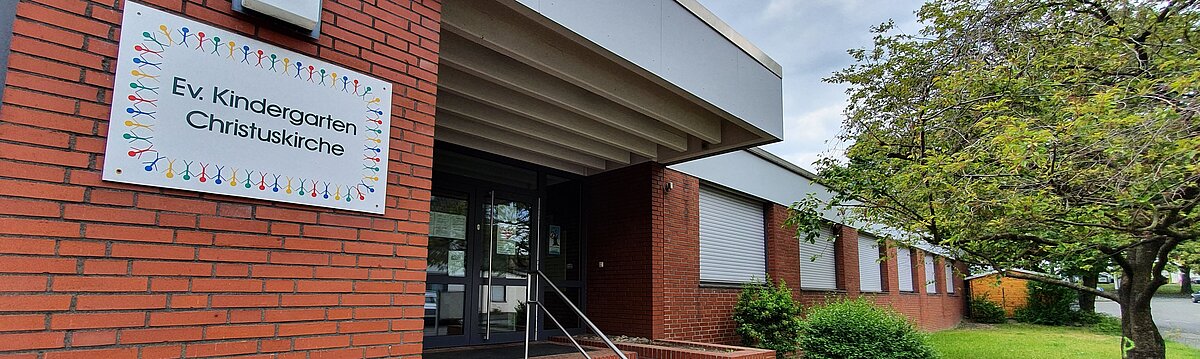 Foto: Eingangsbereich zum Kindergarten an der Sandstraße. In der linken Bildhälfte eine rote Ziegelsteinmaier mit einem großen Schild und dem Schriftzug E. Kindergarten Christuskirche umrahmt von bunten Strichmännchen, in der rechten Bildhälfte funf aufwärtsführende Stufen mit einem Geländer, die zu den Eingangstüren führen, rechts daneben weitere fenster des febäudes mit heruntergelassenenen Rollos