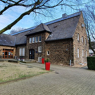 Foto: Eingangsbereich des Gemeindezentrums am Steinhügel: Ein gepflasterter Platz mit einem eingefassten Wiesenbeet um einen großen Baum, mehrere Blumenkübel. Im Hintergrund ein Haus aus Sandstein, Blick auf den Eingangsbereich und die Giebelwand, sowie einen seitlichen Anbau, der ebenfalls Einganstüren ausweist 