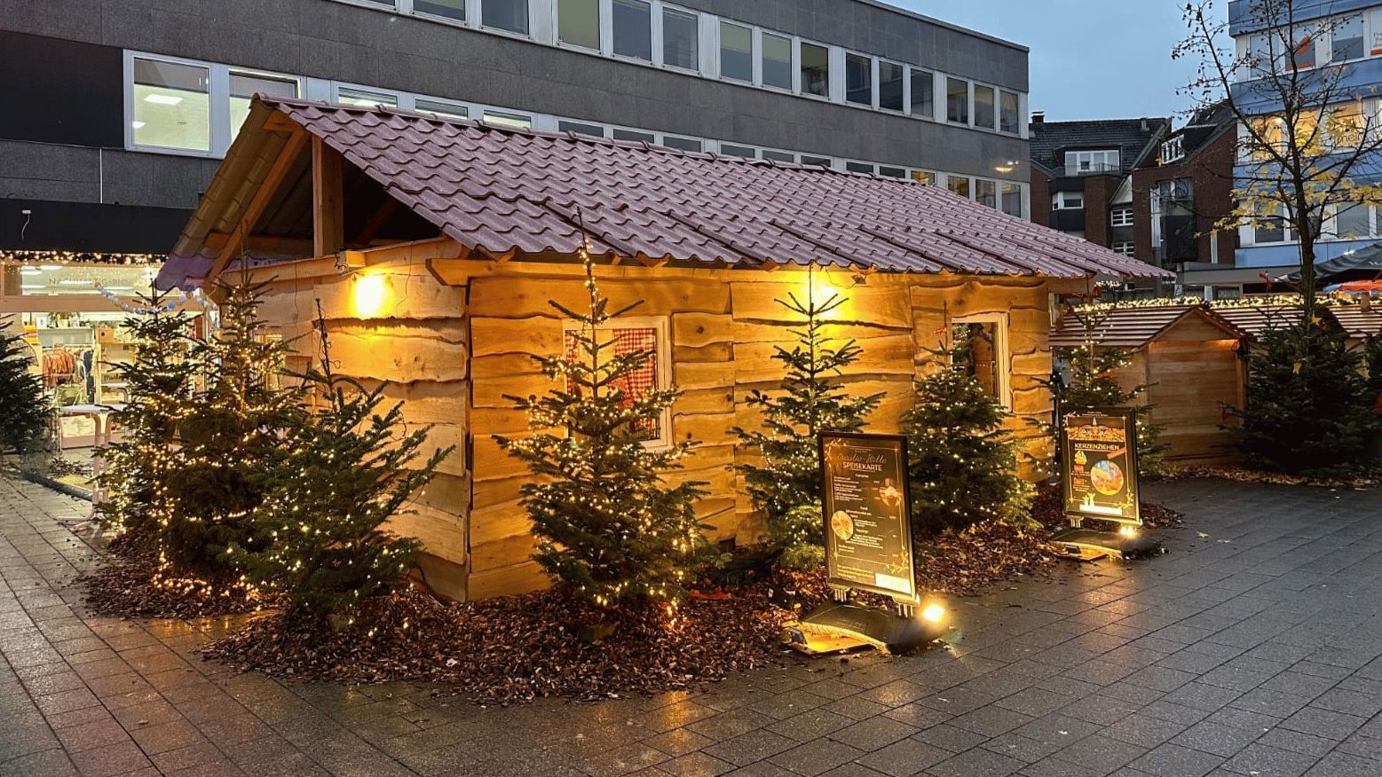 Auf dem Bild ist im Hintergrund die Stadtgalerie in Witten zu sehen. Davor steht die Creativ-Hütte. Sie ist aus Holz und sieht aus wie eine Almhütte. Sie ist bunt beleuchtet. Um sie herum stehen Tannenbäume, die mit Lichterketten geschmückt sind. Am Boden ist Rindenmulch verteilt.